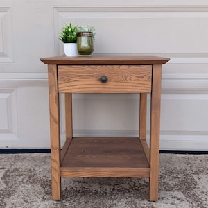 May include: A medium-brown wooden nightstand featuring a single drawer with a small, dark circular knob, a flat top surface, and an open lower shelf. The wood grain is visible throughout the structure, which stands on four straight legs.