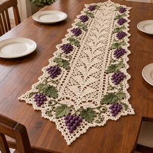 May include: A cream-colored crocheted table runner featuring an intricate openwork lace pattern. The runner is decorated with three-dimensional purple grape clusters and green vine leaves appliqued along the scalloped edges.