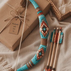 May include: A crocheted toy bow and three arrows featuring a geometric pattern in turquoise, brown, white, and orange. The bow has a string and brown tassels at both ends. The arrows have wooden shafts with crocheted tips and fletching.