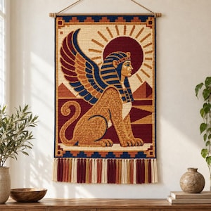 Op de afbeelding: Een rechthoekig wandkleed met een geweven afbeelding van een gevleugelde sfinx in profiel. Het ontwerp bevat een donkerrode zon, blauwe en gouden vleugels en een beige lichaam op een crèmekleurige achtergrond, afgewerkt met een donkerblauwe rand.