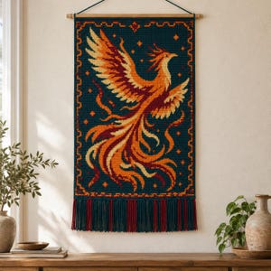 Könnte beinhalten: Ein rechteckiger Wandbehang mit einem Kreuzstich-Phönix in Orange-, Gelb- und Rottönen auf dunkelblaugrünem Hintergrund. Das Motiv ist von einer dekorativen Bordüre umrahmt und schließt unten mit Fransen aus dunkelblaugrünem und rotem Garn ab.