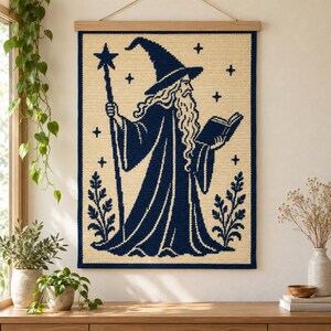 Pode incluir: Uma tapeçaria retangular de crochê apresentando um mago azul-marinho escuro com uma barba longa, um chapéu pontudo e um cajado com uma estrela no topo, segurando um livro aberto. O design está sobre um fundo creme com estrelas e folhagens.