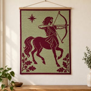 Könnte beinhalten: Ein rechteckiger Wandbehang aus Häkelarbeit mit einem dunkelroten Zentaur-Bogenschützen auf hellgrünem Hintergrund. Das Design zeigt einen Stern in der oberen linken Ecke und Eichenblattmotive in den unteren Ecken, aufgehängt an einer Holzstange.