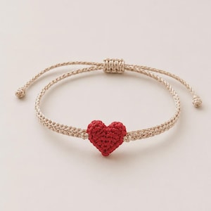 Puede incluir: Una pulsera de cordón trenzado beige con un pequeño corazón rojo de ganchillo texturizado en el centro. La pulsera incluye un cierre deslizante ajustable con dos extremos de cordón finos rematados con pequeños nudos.