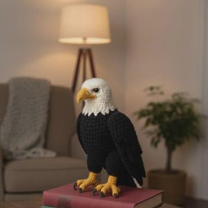Op de afbeelding: Een gehaakt beeldje van een Amerikaanse zeearend met een witte kop, een gele snavel, zwarte lichaamsveren en gele klauwen. De adelaar staat bovenop een stapel van twee boeken. De textuur van het garen is zichtbaar over het hele vogelbeeldje.