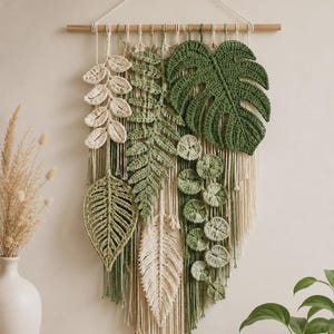 May include: A wall hanging featuring various crocheted botanical elements in shades of cream, sage, and forest green. The piece includes a large monstera leaf, a fern, and circular leaves suspended from a wooden dowel with cream-colored cord.