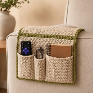 Puede incluir: Un organizador de reposabrazos de ganchillo en color beige con un borde verde oscuro y tres bolsillos. Los bolsillos contienen un mando a distancia negro con botones morados, unas gafas de montura oscura y un cuaderno de espiral marrón.
