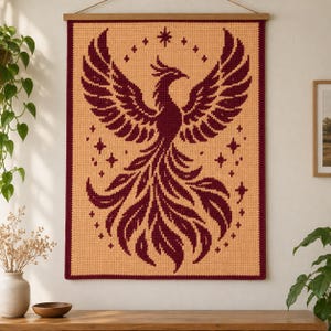 Könnte beinhalten: Ein rechteckiger Wandbehang aus Häkelarbeit mit einem dunkel kastanienbraunen Phönix-Motiv auf hellpfirsichfarbenem Hintergrund. Der Phönix ist mit weit ausgebreiteten Flügeln dargestellt und von kleinen sternförmigen Akzenten umgeben.