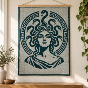Könnte beinhalten: Ein rechteckiger Wandbehang mit einem dunkel-türkisfarbenen Häkeldesign von Medusa auf hellgrauem Hintergrund. Die zentrale Figur ist von einer kreisförmigen griechischen Mäanderbordüre umgeben. Das Textil hängt an einer hölzernen Stange.