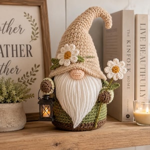 Könnte beinhalten: Eine gehäkelte Gnomenfigur mit einer beigen, spitzen Mütze, die mit einem weißen Gänseblümchen verziert ist, einem langen weißen Garnbart und einem grünen Körper. Der Gnom hält eine kleine schwarze Laterne und ein Gänseblümchen am Stiel.