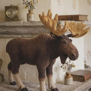 Op de afbeelding: Een handgehaakte elandfiguur met een donkerbruin lichaam, lichtbeige onderbenen en grote, getextureerde tan-kleurige geweien. Het knuffeldier heeft een prominente snuit, een kleine baardachtige pluk onder de kin en donkere, ronde ogen.