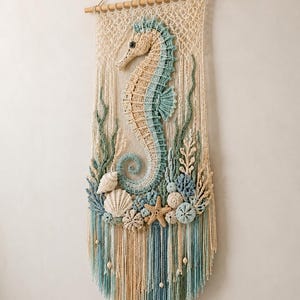 May include: A macrame wall hanging featuring a central crocheted seahorse in beige and teal, surrounded by crocheted seashells, a starfish, and sea plants. The piece is suspended from a wooden dowel with a hanging cord and long fringe.