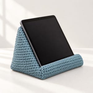 May include: A dusty blue, triangular crochet tablet stand with a textured, yarn-based surface. The stand features a wide, horizontal ledge at the base to securely hold a black tablet in a tilted, upright viewing position.