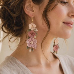 May include: A pair of long, dangling earrings featuring three crocheted flowers in shades of cream, dusty rose, and mauve, accented with small green crocheted leaves. The floral arrangement is suspended from a simple gold-toned metal hook wire.