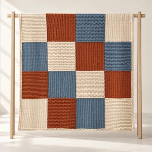 May include: A knitted blanket featuring a four-by-four grid of alternating square patches in cream, terracotta orange, and slate blue. The blanket is displayed hanging on a light-colored wooden frame, showcasing a consistent garter stitch texture throughout the entire piece.