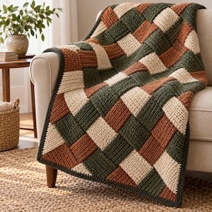 May include: A chunky knit throw blanket featuring a woven basket-weave pattern in alternating squares of cream, forest green, and burnt orange yarn. The blanket is draped over the arm and seat of a neutral-colored upholstered chair.