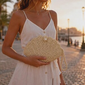 Könnte beinhalten: Eine beige gehäkelte Clutch in Muschelform mit einer einzelnen weißen Perle in der Mitte der Oberkante und einer passenden gehäkelten Handschlaufe, gehalten vor einem weißen ärmellosen Kleid mit durchgehender Knopfleiste auf der Vorderseite.