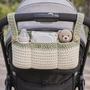 May include: A crochet stroller organizer featuring a sage green and cream color scheme with three compartments. It attaches to the handlebar with two straps secured by wooden buttons. The organizer holds a baby bottle, wipes, and a small teddy bear.