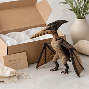 Peut inclure: Une figurine de ptérodactyle crochetée avec un long bec, du fil marron foncé et beige, et des ailes déployées. À côté se trouvent une boîte d'expédition en carton ouverte, une petite carte beige avec le texte thank you handmade with care.
