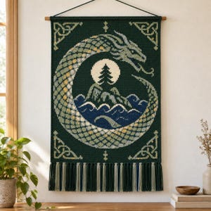 May include: A rectangular dark green wall hanging featuring a crocheted dragon forming a circle around a central scene of a pine tree on a hill above blue waves. The piece includes cream-coloured Celtic corner accents and a long fringe at the bottom.