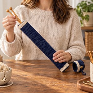 May include: A long, cylindrical, navy blue crochet case with a cream-colored trim at both ends, designed to hold knitting needles. The case is shown with two wooden knitting needles inside and a matching small, round, detachable cap.