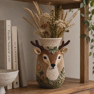 Può includere: Un vaso cilindrico all'uncinetto con un design a forma di muso di cervo color marrone chiaro e crema, palchi marrone scuro, piccole orecchie e dettagli a foglia verde ricamati alla base e sul collo, riempito con un bouquet di grano essiccato.