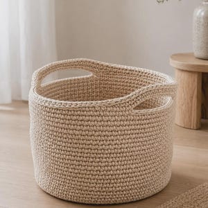 May include: A round, beige, hand-crocheted storage basket made from thick cord. The basket features a textured, dense stitch pattern and two integrated, cutout handles on opposite sides of the rim, providing a sturdy and open design for storage.