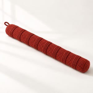 May include: A long, cylindrical, rust-red knitted bolster pillow featuring a textured surface with evenly spaced, raised horizontal bands and a small loop attached to one end for hanging or easy handling of the elongated cushion.