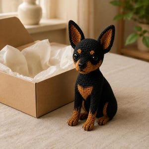 May include: A handcrafted crochet dog figurine featuring black and tan yarn. The small toy has large dark eyes, pointed ears, and a seated posture. It is positioned next to an open cardboard gift box filled with white tissue paper.