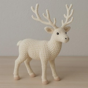 May include: A cream-colored crocheted deer figurine featuring a textured yarn body, large branching antlers, and small dark eyes. The deer stands on four legs with light tan hooves, showcasing detailed stitch work throughout its entire form.