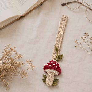 Pode incluir: Um marcador de livros em crochê bege, longo e retangular, com um cogumelo vermelho tridimensional com pontos brancos. O caule do cogumelo é bege e é acentuado com pequenas folhas de crochê verde-escuras na base e perto do topo do caule.