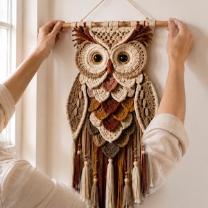 May include: A wall hanging featuring a crocheted owl design in shades of cream, tan, brown, and rust. The owl has large black eyes, layered feather-like crochet details, and a fringe base with wooden beads, suspended from a wooden dowel rod.