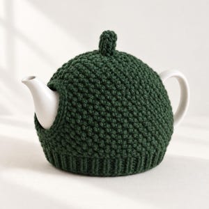 May include: A white ceramic teapot encased in a dark forest green hand-knitted tea cozy. The cozy features a textured moss stitch pattern, a ribbed bottom edge, and a small decorative knob at the top, leaving the spout and handle exposed.