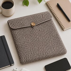 May include: A rectangular, taupe-colored crochet sleeve featuring a textured, dense stitch pattern. The sleeve includes a fold-over flap secured by a single, round, light-brown wooden button centered on the front edge of the flap.