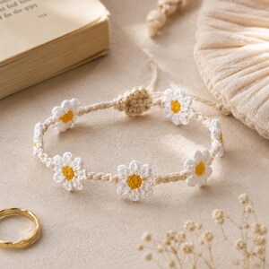 Puede incluir: Una pulsera de ganchillo hecha a mano que presenta una serie de flores de margarita blancas con centros de color amarillo brillante, conectadas por un cordón fino de color beige. Incluye un cierre de nudo corredizo ajustable de hilo beige.