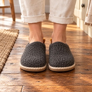 May include: A pair of dark gray crochet slippers featuring a textured knit upper, a light beige braided rope trim around the base, and a small mustard yellow pull tab at the heel, set on a flat, neutral-colored sole.