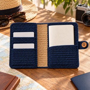 May include: A navy blue and tan crocheted notebook cover featuring two card slots on the left side, a central spine, and a pocket on the right side holding a white notebook. The cover includes a small wooden button closure on the right edge.