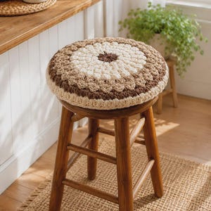 May include: A round wooden stool featuring a thick, circular crochet seat cover. The cover displays a concentric pattern with a dark brown centre, followed by rings of cream, light tan, and dark brown yarn, finished with a cream-coloured edge.