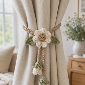 May include: A crochet curtain tieback featuring a central white flower with a tan center, two green leaves, and two dangling stems ending in small white and green bell-shaped flower buds, all attached to a tan braided cord wrapped around a curtain.