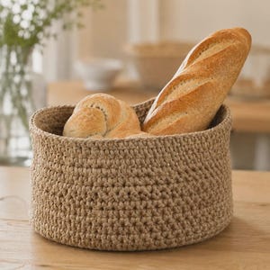May include: A round, beige, crocheted basket with a textured stitch pattern. The basket is filled with a golden-brown baguette and a round, braided loaf of bread, showcasing a contrast between the soft fiber texture and the crusty baked goods.