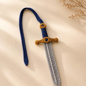 Puede incluir: Un marcapáginas de ganchillo con forma de espada, hoja gris, mango azul oscuro y guarda dorada. El mango y la guarda están decorados con pequeñas cuentas redondas de color azul oscuro. Una larga cola de ganchillo azul oscuro sale del pomo.