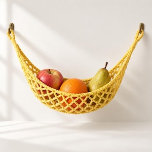 Puede incluir: Una hamaca para frutas tejida a ganchillo en color amarillo brillante, suspendida de dos ganchos de pared color latón. La red contiene una manzana roja, una naranja redonda y una pera verde, como solución de almacenamiento funcional.