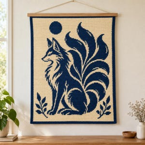 Puede incluir: Un tapiz de pared de ganchillo rectangular que presenta la silueta de un zorro de nueve colas azul oscuro sobre un fondo crema. El diseño incluye un círculo azul oscuro sólido sobre el zorro y pequeñas ramas de hojas en las esquinas inferiores.
