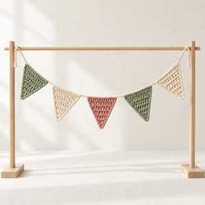 May include: A crochet bunting banner featuring five triangular flags in alternating sage green, cream, and dusty rose colors, suspended from a light-colored wooden frame by a thick, braided off-white cord tied at both ends.