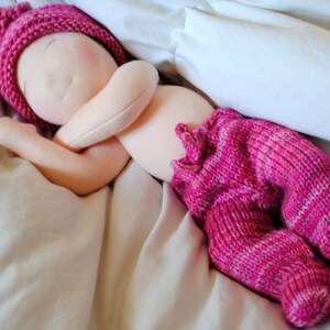 May include: A handmade cloth doll with a pink knitted hat and pink knitted pants, lying on a white blanket.