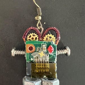 May include: A robot-shaped earring constructed from electronic components. The head is a green circuit board with two gold gears, copper wire coils, a red circular element, and a red crystal. Silver screws form the arms, and two metal nuts form the feet.