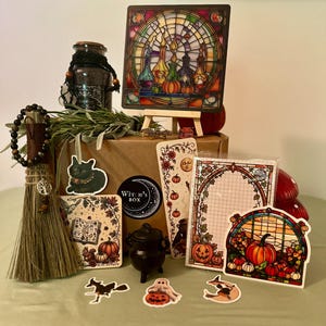 Peut inclure: Un coffret Witch's Box contenant un petit balai avec une breloque arbre, un chaudron noir, une impression d'art style vitrail, un bloc-notes, des autocollants de chat noir, fantôme et citrouilles, et une bouteille en verre avec cordon noir.