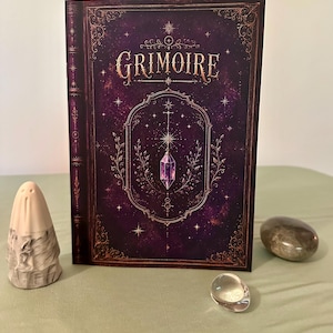 Peut inclure: Un livre rectangulaire intitulé GRIMOIRE avec une couverture violet foncé ornée de filigranes dorés, d'étoiles et d'une illustration de cristal. À gauche, une petite figurine de fantôme blanc cassé et deux pierres polies lisses.