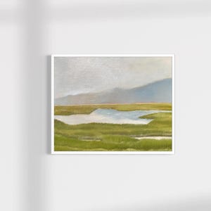 May include: A framed landscape painting featuring a marshy wetland with vibrant green grasses, a central pool of light blue water, and soft, muted blue-grey mountains in the distance under a pale, overcast sky. The artwork is enclosed in a white frame.
