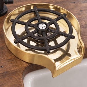 Pode incluir: Um enxaguador de copos em metal dourado com uma grelha circular de plástico preto em forma de leme de navio. O dispositivo possui um bico central para pulverização de água e uma saída larga e curva para drenagem para o lava-loiça.